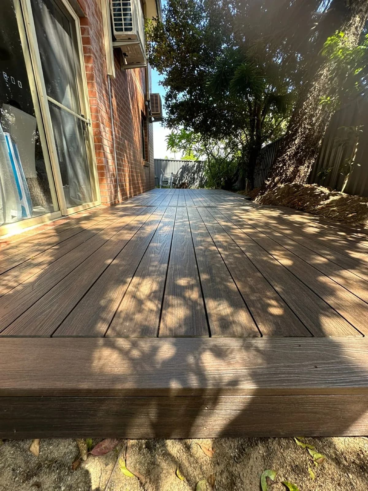Dark brown composite deck along brick wall beside sliding door, fascia edge, dappled light