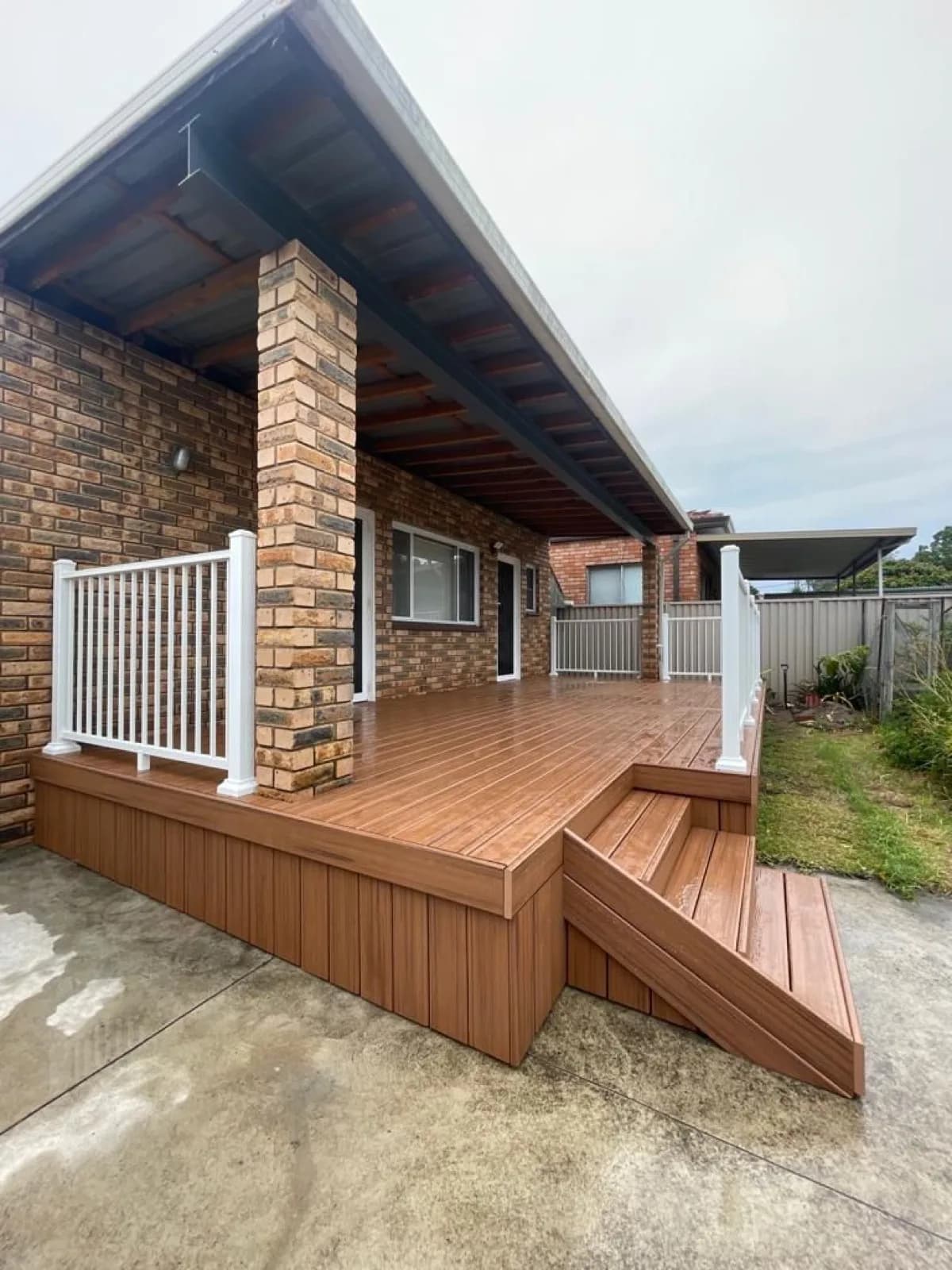 Raised orange-brown composite deck under existing roof with white aluminium balustrade and side steps